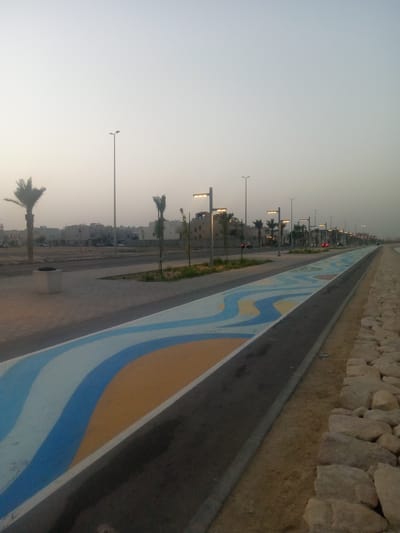 The image depicts a public recreational area in Al Khobar, Saudi Arabia, at dusk. A painted pathway with a wave-like design of blue, white, and yellow curves stretches into the distance. The pathway runs parallel to a road with palm trees and modern streetlights lining its edge. A low barrier of large rocks runs along the right side of the pathway. Buildings can be seen in the background across the road. One person on a motorcycle is visible on the road.