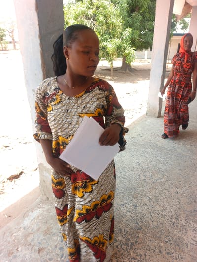 The image captures an outdoor scene in the city of Mada, Nigeria, under a covered walkway. In the foreground, an adult female with dark skin stands, facing slightly right and looking towards her right. She has dark hair styled in a ponytail, wears small gold-colored stud earrings and a thin gold-colored necklace. Her attire consists of a long, patterned dress with a light background featuring dark wavy lines and prominent yellow and reddish-brown floral or abstract designs. She holds a stack of white, lined papers in her right hand, resting against her abdomen. To her right and in the mid-ground, a second adult female walks towards the right side of the frame, slightly away from the camera. She wears a long, loose-fitting dress with an orange and dark patterned fabric, a red head covering, and black sandals. Her right hand is down, appearing to hold a small, dark object. Both individuals are on a light-colored terrazzo or concrete floor. A light-colored rectangular pillar is visible to the left of the foreground subject, and another pillar is further back on the right. Beyond the covered area, a dirt ground is visible, with mature trees featuring green foliage, and light-colored building structures in the distance under bright daylight.