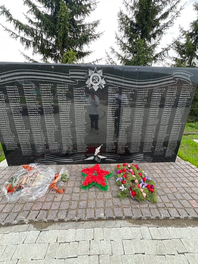 Here is a description of the image:
**Scene:** The image depicts a memorial monument in Введенская Слобода, Russia, likely a war memorial. The memorial is a large, black granite slab, standing vertically. Engraved on the monument are names in Cyrillic script, organized in columns, likely commemorating individuals. The monument is constructed of granite blocks.
**Objects:**
* Three floral wreaths are placed on the cobblestone ground in front of the monument. One wreath is arranged with red and green flowers in the shape of a Soviet star, another is a traditional circular wreath with assorted colorful flowers, and the third is wrapped in cellophane with an orange and black ribbon.
**Environment:**
* The monument is situated outdoors, next to a sidewalk or paved area constructed of grey cobblestones.
* Green foliage from nearby trees is visible in the background, suggesting a park-like setting.
**People and Actions:**
* A reflection of a person taking the photo is visible on the monument's surface. No other individuals are directly visible.