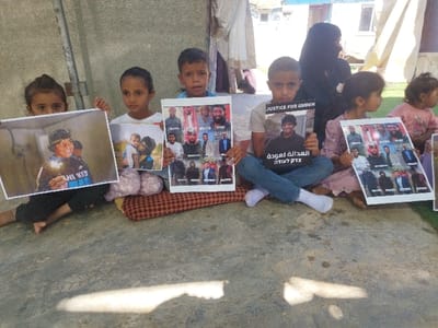 The image displays an outdoor scene in Carmel, Palestinian Territory, featuring five children prominently seated on a concrete surface, holding various printed materials. From left to right: a young girl holds a horizontal photograph of a boy holding a sparkler; a second young girl holds a vertical photograph depicting an adult holding an infant; a young boy in a light blue shirt is positioned centrally; a fourth child, a boy wearing a grey t-shirt and blue jeans, sits cross-legged and holds a vertical sign with a portrait of a young man, bearing the English text "JUSTICE FOR AWDEH" and additional text in Arabic and Hebrew; a fifth child, a young girl in a pink dress, holds a vertical collage of nine male portrait photographs. Another child, partially visible on the far right, holds a similar collage. An adult female wearing a black niqab and abaya is partially visible behind the children on the right. The children are positioned on reddish-brown patterned cushions. The background includes a light-colored concrete wall on the left and temporary structures, resembling tents with light fabric and blue tarpaulin, on the right. Green ground cover is visible on the far right. The scene is illuminated by daylight, creating shadows on the concrete surface.