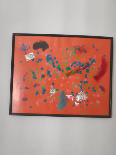 A framed artwork featuring an orange background with various abstract elements is displayed on a white, textured wall. The artwork is rectangular with a black frame. The abstract elements include splatters of blue, green, and pink paint, as well as some glittery blue and silver shapes. There are also a few feathers, a piece of black netting with a honeycomb pattern, and a cluster of small white beads. The overall impression is one of a vibrant and eclectic collage. The location is the city of Eitorf, Germany.