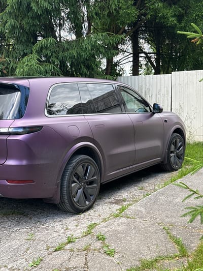 A matte purple electric SUV is parked on a cracked concrete driveway next to a white wooden fence and tall green trees. The SUV has black rims and tinted windows, and the reflection of buildings and trees can be seen in the windows. The driveway is surrounded by green grass and some weeds are growing through the cracks in the concrete. The scene appears to be in a residential area in Kommolovo, Russia. There are no people visible in the image.