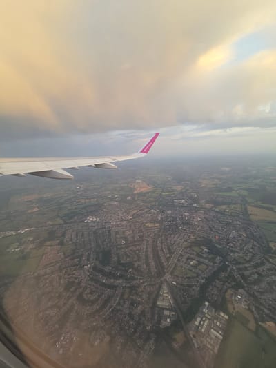 The image shows an aerial view from an airplane window, with a Wizz Air wing visible on the left. The landscape below is a mix of urban and rural areas, including fields and dense residential areas, possibly in the city of St Albans, United Kingdom. The sky is cloudy.