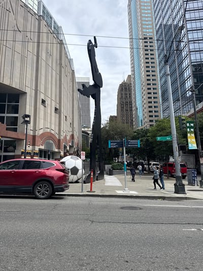 This image depicts a street scene in Seattle, Washington, USA, featuring a large, abstract black sculpture in the foreground, resembling a figure in motion. To the left of the sculpture is a building with a textured, light-colored facade and arched entrances. In front of this building and near the sculpture, a large, inflated white and black soccer ball is visible. A red SUV is parked on the street to the left of the soccer ball.
Behind the sculpture and building, a cluster of tall, modern buildings dominates the skyline, characterized by their glass facades and varied architectural styles. The sky is overcast. Sidewalks are present, with a few pedestrians walking. Street signs are visible, including one indicating "1st Ave." and other directional signs for various locations within the city, such as "Pike Place Market," "Seattle Center Monorail," and "South Lake Union Streetcar." Traffic lights and street lamps are also present. The overall environment is urban, with a mix of contemporary and older architectural elements.