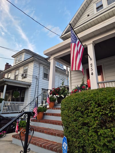 The image shows the exterior of a residential property in South River, United States of America. The property features a two-story house with white siding and a pitched roof. An American flag is prominently displayed on a flagpole extending from the porch. The porch has white columns and a railing. Red brick steps lead up to the porch, flanked by potted plants and smaller American flags. A dark green bush is in the foreground. The house number, "254", is visible on one of the porch columns. A security sign for ADT is partially visible at the base of the bush. The sky above is blue with scattered white clouds. Another house is visible to the left of the main house.