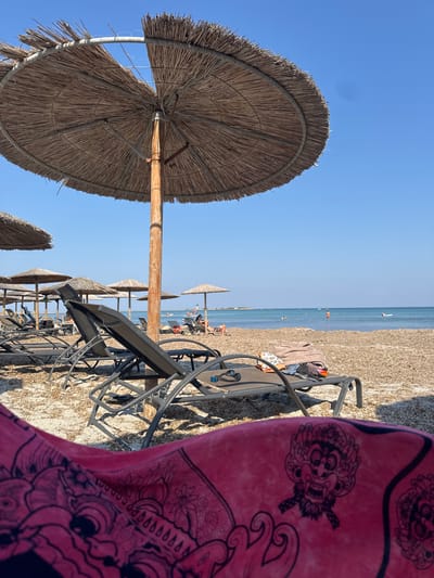 This image depicts a sunny day at a beach with several straw umbrellas and empty lounge chairs arranged on the sand. In the foreground, a large straw umbrella is prominent, casting a shadow. To the right, a pink towel with a black tribal-like pattern lies on top of a black lounge chair, also in the foreground. In the background, the clear blue sea stretches to the horizon, with a few distant figures visible in the water and on the beach. The beach itself appears to be covered in small pebbles and sand. The overall scene suggests a relaxed atmosphere on a warm day, likely in the afternoon given the angle of the shadows. The location is hinted to be Moudros Municipal Unit, Greece. No visible text is present in the image.