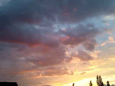 This is a landscape photograph of a dramatic sky at sunset over Mikhaylovka, Kyrgyzstan. The subjects are the clouds and the sky itself, with a hint of a building in the lower-left corner and several trees silhouetted against the bright lower sky. The event taking place is the natural phenomenon of sunset.
Notable details include the time of day, which is clearly sunset, indicated by the warm, golden light illuminating the lower clouds and the horizon. The upper sky is filled with dark, moody clouds tinged with pink and purple hues, suggesting a possibly turbulent weather front or simply a vibrant sunset. The overall mood evoked is one of awe and natural beauty, with a sense of grandeur from the dramatic sky. The location cues of trees and the silhouette of a building anchor the scene to a terrestrial setting. There is no visible text in the image.