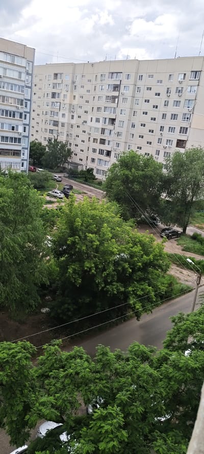 The image depicts a daytime view of a residential area in Syzran, Russia, characterized by multi-story apartment buildings, trees, and parked cars. The sky is overcast with clouds.
In the background, two large apartment buildings are visible. The building on the left is lighter in color and appears to be of a different architectural style than the larger, beige apartment building dominating the center and right of the frame. This larger building has a uniform facade with numerous windows, balconies, and air conditioning units. Antennae and other roof structures are present.
In the midground, a paved road or driveway winds through the scene. Several cars are parked along this road and in adjacent unpaved areas. These include a silver SUV, a white sedan, a dark sedan, and what appear to be other vehicles partially obscured by trees. A street lamp is visible near the road.
The foreground and midground are filled with lush green trees and foliage. The trees are dense, with some appearing to be deciduous. Some of the foliage is so thick that it obscures parts of the parked cars and the ground below.
The overall impression is of a typical urban residential environment during daytime, possibly in the spring or summer due to the green foliage. There are no people visible in the image, and no discernible actions or interactions are occurring.