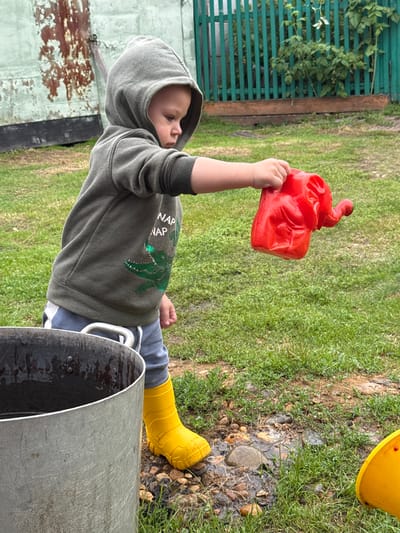 In this image, a young child is depicted outdoors in what appears to be a backyard or garden area in Верхняя Уря, Russia. The child, wearing a grey hooded sweatshirt with a green alligator graphic and the words "NAP NAP", light blue pants, and yellow rubber boots, is holding a red plastic elephant-shaped watering can. Water is visibly dripping from the watering can, suggesting the child is playing with it, possibly pouring water onto the ground.
In the foreground, to the left of the child, is a large, weathered metal pot or cauldron with handles. The ground around the child is covered in grass and scattered stones, with some areas appearing wet, indicating recent moisture. In the background, there is a rustic green wooden fence and a portion of a weathered building with peeling paint. The overall environment suggests a casual, outdoor setting, likely a residential area given the presence of a fence and building.