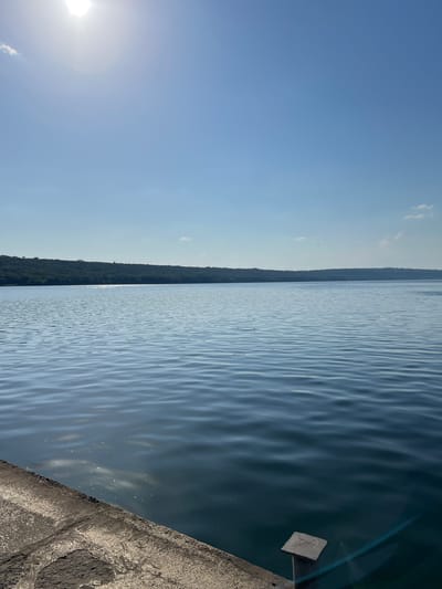 The image shows a body of water with ripples, likely a lake or river, under a clear blue sky. The sun is visible in the upper left corner, suggesting it is daytime. On the right side of the image, there is a concrete embankment or wall with some texture. In the distance, a treeline or forested area is visible along the horizon, separating the water from the sky. The overall scene is serene and appears to be an outdoor landscape. There is no visible text in the image. The orientation of the image is sideways, with the horizon running vertically.