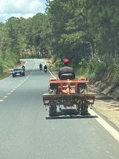 A man wearing a red cap and a black vest with the "DIOR" logo is operating a small, orange tractor on a paved road in Thôn 6, Vietnam. The tractor has a tilling attachment on the back. The road is winding through a forested area with tall pine trees. There are other vehicles on the road, including a black car and several motorcycles. The sky is partly cloudy.