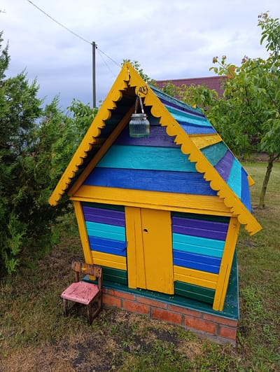 A colorful, miniature house-like structure, possibly a birdhouse or a decorative garden feature, stands in a grassy outdoor setting. The structure is painted in horizontal stripes of yellow, blue, turquoise, and purple, with a bright yellow, scalloped trim around its triangular roof. A clear glass jar with a metal lid hangs from a hook on the front of the roof, serving as a decorative element.
In front of the structure, a small, weathered wooden chair with a faded pink seat is placed on the grass. The house itself is raised on a base made of red bricks. The scene is surrounded by lush green foliage, including a conifer tree on the left and leafy trees in the background. In the distance, a red-tiled roof of a building is visible. Overhead, a utility pole and power lines are present against a cloudy sky, suggesting an overcast day. The overall impression is one of a whimsical, slightly rustic garden decoration. There is no visible text in the image. The location context indicates it is within the Belavushski Sel'ski Savet, Belarus.