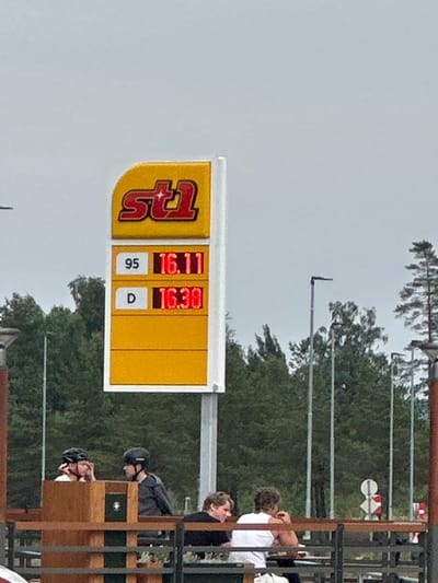 The image shows a close-up of a gas price sign in Slitu, Norway. The sign displays prices for two types of fuel: "95" at 16.11 and "D" at 16.38. The logo of "st1" is prominently displayed at the top of the sign. In the background, a few people are visible sitting at a table. The sky is overcast. The scene appears to be outdoors, likely at a gas station.