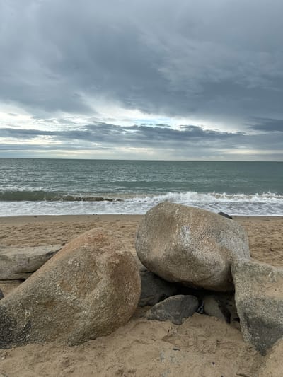 The image depicts a coastal scene at Courtown, Ireland, under an overcast sky. The foreground is dominated by large, weathered boulders resting on a sandy beach. The sand appears damp, suggesting recent wave activity or high tide. In the midground, the sea stretches out, with moderate waves breaking onto the shore, creating white foam. The water is a muted green-blue color. The horizon is clearly defined, separating the sea from the sky. The sky itself is filled with heavy, grey clouds, with lighter patches suggesting breaks in the cloud cover. The overall atmosphere is somber and natural, characteristic of a cloudy day by the sea. There are no people or animals visible in the frame.