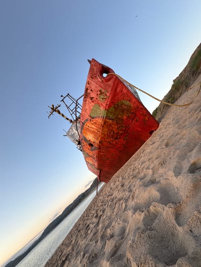 An abandoned red ship is beached on the sandy shore of the Barents Sea in Teriberka, Russia. The ship, heavily rusted and partially covered in green algae, lists to its port side. A thick rope is attached to the ship and stretches across the sand. The sky is a clear, pale blue, with a faint haze near the horizon where the sea meets the distant, low-lying mountains. The sand is textured with visible footprints and wave-like patterns. The low angle of the sun suggests it is either early morning or late afternoon, casting soft light on the scene. The overall atmosphere is one of decay and isolation.