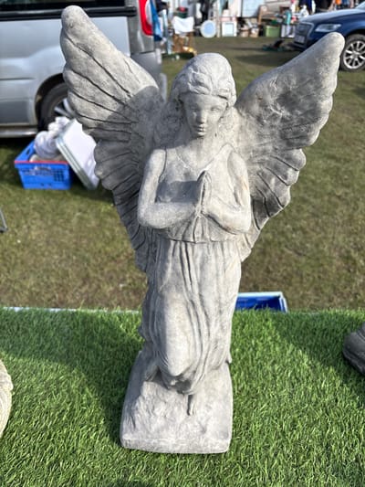 A statue of an angel with large wings stands on a rectangular base. It is made of gray stone and appears weathered. The statue is positioned on a patch of artificial green turf. To the left, out-of-focus, are parts of a vehicle and various items, suggesting a possible market or sale environment. In the background, more grass and another vehicle are visible. The setting is likely an outdoor space in Chirk, United Kingdom.