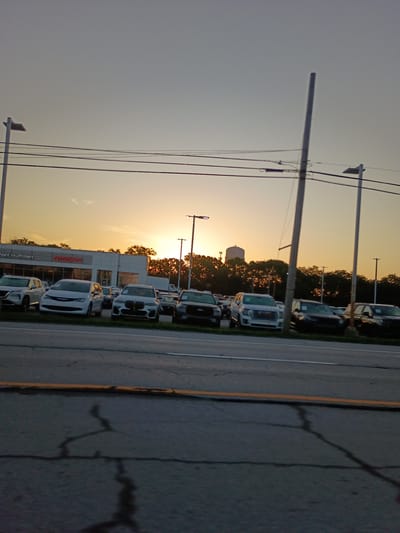 This image captures a car dealership with a row of vehicles parked in front of a building. The sun is setting, casting a warm glow across the sky and illuminating the scene with a golden light. Trees line the background, and a water tower is visible in the distance. The sky is clear, suggesting pleasant weather. A utility pole with wires stands tall in the foreground, adding to the urban setting. The cars appear to be new models, with one prominently displaying the "Nissan" logo. The name "Neil Huffman" is also visible on the building, likely indicating the dealership's name. The overall mood is serene, with the sunset creating a peaceful atmosphere.