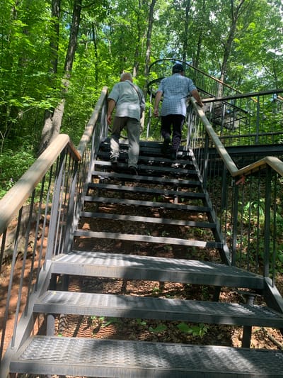 Two men are ascending a metal staircase in a dense forest. The man in the foreground is wearing a light-colored short-sleeved shirt and light-colored trousers, with a strap across his chest. The man behind him is wearing a blue and white plaid shirt, dark trousers with white stripes, and a dark baseball cap. The staircase is made of metal grating and has metal railings with wooden handrails. The forest floor is covered with fallen leaves and some green undergrowth. Sunlight filters through the trees, casting dappled shadows on the stairs and the ground. The image appears to be taken in Arbuzov-Baran, Russia, based on the provided context.