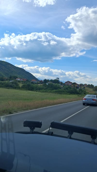 The image depicts a scenic drive through a rural area, likely on the outskirts of Novo Leski, Bulgaria. The foreground shows the hood of a vehicle with two auxiliary lights mounted on it. Beyond the vehicle, a two-lane asphalt road stretches into the distance, marked with a white dashed line.
On the right side of the road, a silver sedan is driving away from the camera. The car has a license plate that appears to read "CB175 AO".
The landscape transitions into a rolling hillside covered in dry grass and scattered bushes. Further back, a cluster of houses with red-tiled roofs are nestled amidst green trees, suggesting a small village. The background is dominated by a large, verdant mountain that slopes down towards the village. In the far distance, snow-capped mountains can be seen, adding a dramatic contrast to the green landscape.
The sky is a vibrant blue, punctuated by dramatic, fluffy white clouds, indicating fair weather. The overall impression is one of a peaceful and picturesque rural journey.