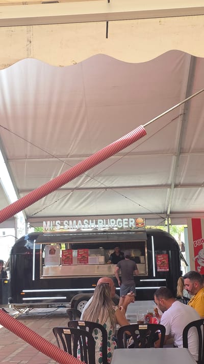 This image shows an outdoor scene at what appears to be a food truck festival or market under a large white tent. The central focus is a black food truck with the name "MRS SMASHBURGER" written vertically in illuminated white letters. The truck has a serving window where a staff member is visible behind the counter, and a customer is interacting with them. Several people are seated at tables in front of the food truck, seemingly eating and socializing. One person in the foreground is facing away from the camera, with long blonde hair. Another man is looking down at food on his plate, and a third man is leaning over. The overall atmosphere seems casual and relaxed.
Notable details include the bright daytime lighting, suggesting it is likely midday. The setting under a tent indicates an outdoor event, possibly in a town square or a designated market area. The red corrugated tubes hanging from the tent structure are a distinctive visual element. The image is oriented vertically, suggesting it was taken with a phone camera in portrait mode.
Visible text includes "MRS SMASHBURGER" on the food truck and "BEST" printed on several posters displayed on the truck's exterior, likely advertising menu items or specials. A red sign with white lettering is partially visible at the top of the frame, reading "FOR".