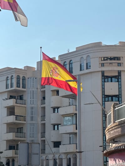 The image shows the Spanish flag waving against a bright blue sky, with a modern building in the background. The flag is predominantly red and yellow, with the Spanish coat of arms visible on the yellow stripe. The building has a contemporary architectural style, featuring a light-colored facade with numerous windows, some of which are set in arched recesses. The flag is attached to a flagpole that extends from the building. Another flag, partially visible in the lower left corner of the image, appears to be white and red. The scene suggests an outdoor setting on a clear day, likely in Motril, Spain, given the context. There is no discernible activity or event taking place, other than the flags flying. Visible text on a sign at the top right reads "bienvenido a la mesa". The time of day appears to be daytime, with the clear sky and strong lighting. There are no people visible in the image.