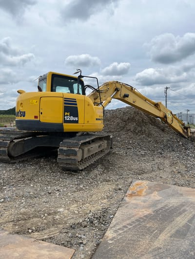 An excavator, identified by the "KOMATSU PC 128US" marking on its yellow body, is positioned on a construction site. The excavator is surrounded by gravel and dirt. A large pile of gravel is located behind the excavator, with its boom extending over it. The sky above is overcast with clouds. In the background, utility poles and industrial buildings are visible, suggesting an urban or suburban setting. The ground in the foreground is covered with gravel and shows tire tracks from heavy machinery. A metal plate with tire marks is also present in the foreground. The image does not contain any people. The scene is consistent with a construction area in Hanno, Japan.