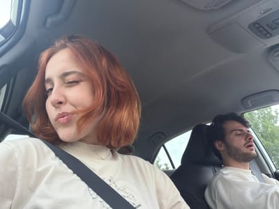 The image is taken inside a car, likely a selfie, showing two individuals wearing seatbelts. A woman with red hair is in the passenger seat, winking at the camera with a pursed lip expression. A man with dark hair and a beard is in the driver's seat, looking towards the sky with his mouth slightly open. The car appears to be moving, indicated by trees visible through the car windows. The interior of the car is modern with a light gray ceiling and dark gray seats. The location is unknown, as the city of Mrgvali Chala, Georgia cannot be confirmed from this photograph.