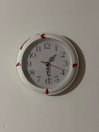 A white, circular analog clock with Roman numerals is mounted on a neutral-colored wall. The clock face is white with black numerals and a red second hand. The hour and minute hands are black. The clock's frame has a distressed, white finish with red paint splatters. The wall is slightly textured and shows minor imperfections. There is a shadow cast by the clock on the wall to its right. The image appears to be taken indoors, possibly in a residential setting within Радваніцкі сельскі Савет, Belarus. No people are present in the image.