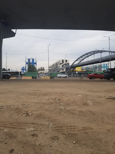 The image depicts an overcast day in a city, likely Бұлақты, Kazakhstan, with construction or undeveloped land in the foreground. A large, dark, overhanging structure, possibly a bridge or overpass, dominates the upper portion of the frame. Beyond this, a street runs horizontally, with various vehicles visible.
In the mid-ground, several green buses and a dark-colored truck with visible wheels are present on the street. A red sedan is also parked on the right side of the street. Behind the street, a pedestrian bridge with a distinctive arched structure arches across the scene. Buildings with modern architecture, some featuring large blue and white cross designs, are visible in the background. Streetlights and power lines are also interspersed throughout the scene. The foreground consists of dry, dusty soil with tire tracks and scattered debris, suggesting an area undergoing or awaiting development. No people are visible in the image.