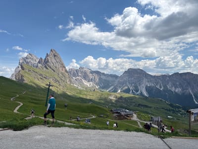 This is a landscape photograph taken on a sunny day with scattered clouds, likely in the late morning or early afternoon. The scene depicts a mountainous region with lush green meadows dotted with yellow wildflowers. A winding dirt path leads through the landscape, with several people walking along it. In the midground, a cluster of buildings forms a small village or mountain resort, with a yellow excavator indicating ongoing construction.
The prominent subjects are the mountains, characterized by their rugged, rocky peaks and steep slopes, some of which are covered in scree. The sky is a vibrant blue, with fluffy white clouds casting shadows on the mountains and the land below. Several ski lifts are visible, suggesting the location is also a ski resort during the winter months.
Several individuals are present in the image, walking along the paths and enjoying the scenery. One person in the foreground, a man wearing a teal shirt and dark leggings, is looking out towards the mountains. Another person, a woman in a dark top and shorts, is walking up a path behind him. Other figures are scattered further down the path and near the buildings.
The overall mood is one of outdoor recreation and appreciation of natural beauty. The location appears to be a popular tourist destination, likely in the Dolomites region of Italy, given the context. There is a signpost visible on the right side of the image, but the text is too small to be read.