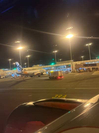 The image shows an airport tarmac at night. Several airplanes are visible, including a Jetstar aircraft with the registration VH-VGY. A baggage tug labeled "ONATA" is also present. In the foreground, the wing of another aircraft, likely the one from which the photo was taken, is visible. Airport lights illuminate the scene. There are no visible people in the image. The location is an airport in Moss Vale, Australia.