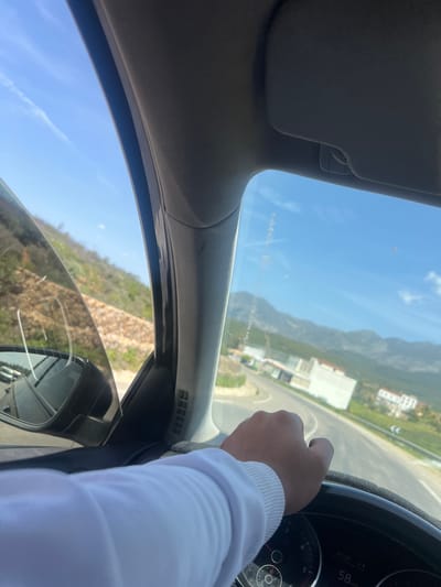 The image, taken from inside a car, shows a person driving along a road in Derdara, Morocco. The driver's hand is visible on the steering wheel. The speedometer shows the car's speed at 58 km/h. Outside the car, a mountain range is visible in the distance, along with buildings. The sky is blue with scattered clouds. The car's side mirror reflects the surrounding environment, including trees. The sun visor is down on the driver's side.