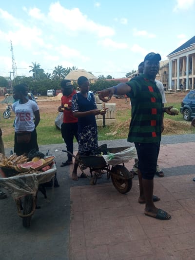 The image portrays an outdoor scene in Ajalli, Nigeria, under a partly cloudy sky. The foreground consists of a reddish-brown patterned paved walkway transitioning to an unpaved, dusty area.
Two wheelbarrows are present. The left wheelbarrow contains various fruits, including sliced watermelon and bananas, partially covered by a white cloth. The right wheelbarrow is mostly empty, containing a white sack.
Four primary individuals are visible. In the foreground right, a male wearing a dark green and red striped polo shirt, dark shorts, dark slide sandals, and a dark baseball cap holds a pale green fruit in his right hand while extending his left index finger. To his immediate left and slightly behind, a female wearing a dark blue dress with a white floral pattern, barefoot, holds a similar pale green fruit. Behind her, a male in a red t-shirt, black pants, and a dark baseball cap with "NYPD" lettering holds a yellow fruit. Further left, a female wearing a white t-shirt with "Say YES" printed on it and dark lower attire, with a pouch around her waist, stands facing left. Other indistinct figures are visible in the background.
The background features multiple structures. On the far right, a large light-colored multi-story building with classical-style columns and balconies is prominent. Other less distinct buildings are visible further back. Vehicles include a motorcycle on the far left, a white van in the mid-ground, and a dark blue SUV parked on the far right. Piles of concrete blocks are present in the mid-ground. A tall metal communication tower is visible on the far left, and palm trees along with other vegetation are scattered throughout the background.