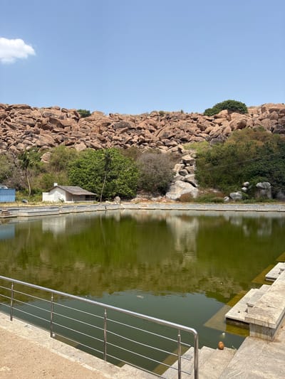 The image depicts a rock formation adjacent to a body of water, possibly a pond or lake, in what appears to be a rural or natural setting. The rock formation is primarily composed of large, rounded boulders, and sparse vegetation grows among them. A small, white, building with a simple roof structure is situated near the water's edge, nestling among the trees.

The water has a green tint. A concrete structure with steps leading into the water, indicating the pond is man-made, is visible along the side. A silver railing runs along the top of the structure. 

The sky is a clear, pale blue, suggesting a sunny day.