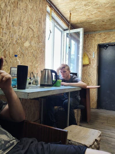 The photograph captures an indoor scene, possibly a rustic dwelling or temporary structure, with walls constructed from oriented strand board (OSB) panels. A young man, appearing to be of Caucasian descent and in his late teens or early twenties, is seated at a table, his right hand resting on his forehead, suggesting contemplation or weariness. He is wearing a grey t-shirt and dark trousers.

In front of him on the table are several objects, including a silver electric kettle with the brand "wiron" visible, a black tall cup, a green bowl, and a glass jar containing various utensils like spoons and forks. A large plastic bottle of water with a blue cap is also present on the table. To the left of the frame, a person's arm and hand are visible, with the hand held up, possibly indicating a gesture.

The environment features a large window to the left, allowing natural light to illuminate the room. A yellow bag hangs from the ceiling near the window. The floor is made of wooden planks. The overall impression is one of a simple, utilitarian space. The image was taken in the city of Vilne, Ukraine.