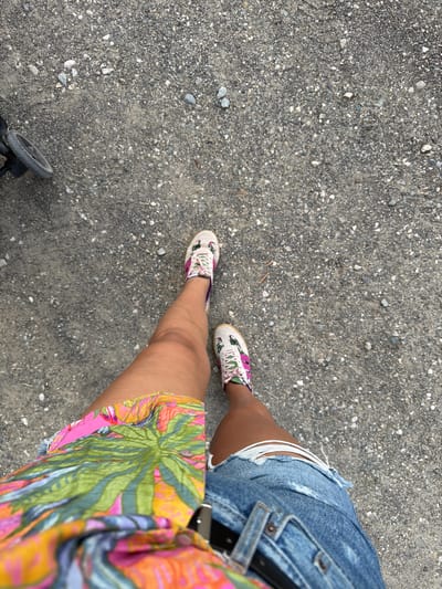 The image shows a person's legs and feet from a top-down perspective, standing on a gravel surface. The person is wearing denim shorts with frayed edges and colorful sneakers with a floral pattern. A brightly colored garment with a tropical leaf print is visible on the right side of the frame, draped over the person's leg. A small portion of a stroller wheel is visible in the bottom left corner of the image. The scene suggests an outdoor setting, possibly a park or a walkway, on a clear day. No text is visible in the image.