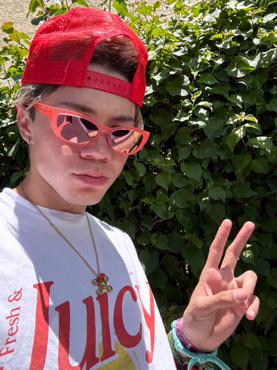 The image depicts a person wearing a red mesh cap and pink sunglasses, standing in front of a green leafy background. The person is making a hand gesture with their right hand, showing three fingers extended. The person is also wearing a white t-shirt with a red design and a gold necklace. The image appears to be taken outdoors during daylight. Based on the presence of lush green foliage, the location is likely in Indian Wells, United States of America, a city known for its landscaping and resorts.