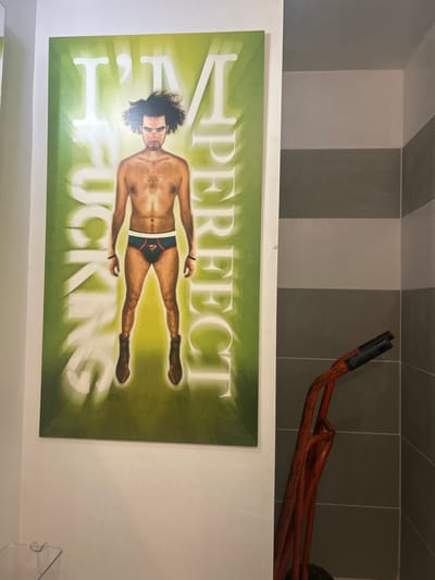 The image depicts an indoor setting in Cucuron, France. A large, vertically oriented poster is mounted on a plain white wall on the left side of the frame. The poster features a shirtless male figure, appearing to be of adult age, with dark, frizzy hair and an intense facial expression, looking directly forward. He is wearing dark briefs with a red and yellow Superman 'S' shield logo on the front and dark brown boots. White capital letters frame the figure, spelling "I'M FUCKING PERFECT" vertically on the poster's green background, which has a bright central glow. A faint signature, "Eli Gindi," is visible at the bottom center of the poster. To the right, a corner wall is visible, tiled with alternating horizontal stripes of light grey and dark grey. Leaning against this tiled wall is the upper portion of an orange-red metal hand truck, equipped with black rubberized handles. A small, translucent plastic container is partially visible in the bottom-left foreground.