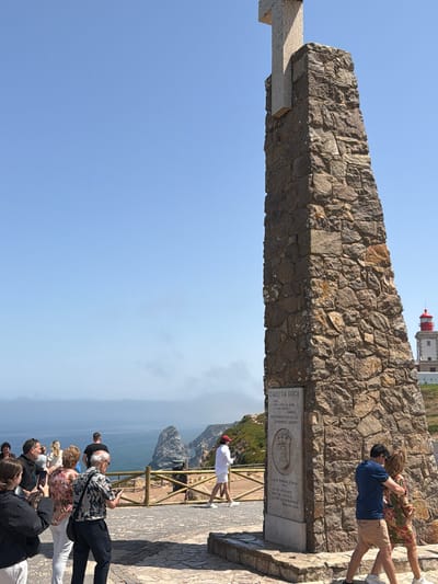 A sunny day at Cabo da Roca, Portugal, the westernmost point of mainland Europe in the municipality of Colares. A large stone monument topped with a cross stands prominently in the foreground. Inscriptions on the monument's base read "CABO DA ROCA" and other details in Portuguese. Several tourists are present, with some posing for photos near the monument, while others are viewed from behind looking out at the ocean. A couple is seen walking away from the monument, the man with his arm around the woman. In the background, the vast Atlantic Ocean stretches to the horizon, with cliffs and a distant lighthouse visible.