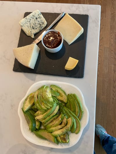 The image shows a plate of sliced avocado on a white marble countertop. The avocado slices are arranged in a circular pattern, with the tips of the slices facing outward. There are also four different kinds of cheese on a black slate platter to the left of the avocado. The cheese includes a blue cheese, two wedges of provolone, and a smaller wedge of cheddar. There is also a small white bowl with a dark brown sauce, likely a balsamic glaze, in the center of the platter. The scene appears to be in a kitchen, with a wooden floor visible in the background. The location based on the coordinates is Rye, New York.