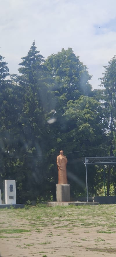 The image captures an outdoor scene with a statue as the central subject. The statue, made of a copper-colored material, depicts a figure in a long robe, standing with one hand held to its chest. It is positioned on a concrete pedestal. To the left of the statue, a white rectangular monument featuring a portrait of a man is visible. In the background, a dense line of green trees, including coniferous and deciduous varieties, forms a natural backdrop. Above the trees, a cloudy sky with some blue patches is present. The foreground consists of a paved area with sparse grass growing between the paving stones. A metal structure with a roof, possibly a canopy or shelter, is situated to the right of the statue, partially obscuring some of the trees. The overall setting appears to be a public space, possibly a park or a memorial site, in Kolomak, Ukraine.