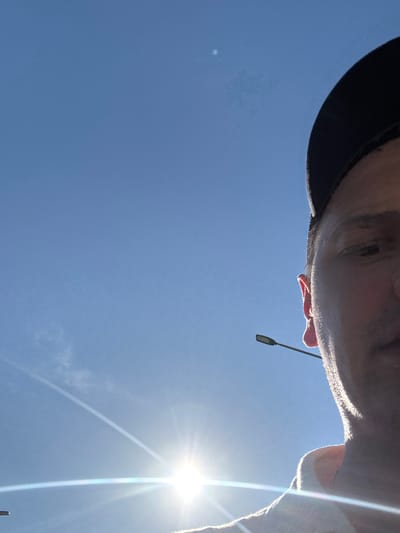 The image is a low-angle, upside-down selfie taken outdoors against a bright blue sky. The sun is shining intensely, causing lens flare and a bright halo effect. A person's face is partially visible at the top of the frame, wearing a dark cap. The person appears to be looking upwards. A small, dark object, possibly a light fixture, is suspended from a thin wire or cable. The scene suggests a sunny day, likely in the afternoon given the sun's position and brightness. There are no visible text elements in the image. The context provided, Koltsovo, Russia, offers a geographical location for the image.