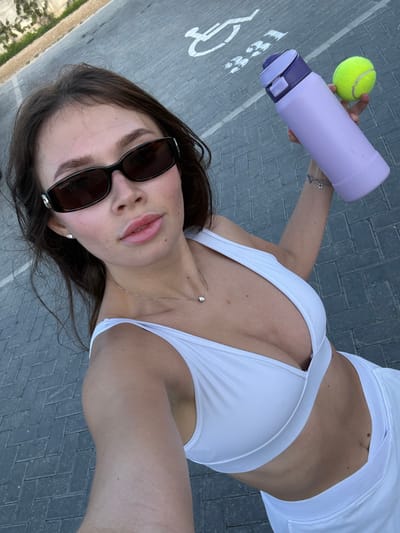 A woman wearing a white sports bra and shorts stands on a brick-paved surface. She is holding a yellow tennis ball and a lavender water bottle in her left hand. She wears dark sunglasses and a silver necklace with a pendant. A partially visible disabled parking symbol is on the ground near her left arm. The location appears to be outdoors, likely in Tserkezoi, Cyprus.
