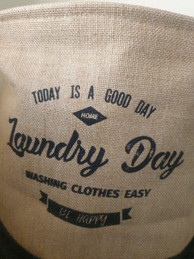 A close-up image depicts a beige, coarse-woven fabric container, likely a laundry hamper or bag, with various black text inscriptions. The top inscription reads "TODAY IS A GOOD DAY". Below this, a small diamond graphic contains the word "HOME". Prominently displayed in a script font are the words "Laundry Day". Further below, the text "WASHING CLOTHES EASY" is visible. At the bottom, a banner graphic contains the words "BE HAPPY" in a script font. The fabric exhibits a visible weave pattern and slight creasing. No people, actions, or direct environmental context beyond the object itself are depicted. The scene is located in Mumbuni North ward, Kenya.