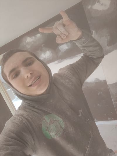This image depicts a young adult male in an interior room located in Deerlijk, Belgium, which appears to be under construction or renovation. The male subject, with light skin and short brown hair, faces the camera directly, displaying a slight smile with his mouth open, revealing teeth. His face is coated with a fine white powder. He is wearing a grey hooded sweatshirt with the hood up; the sweatshirt is heavily covered in white dust and features a circular green logo with a cracked design on the chest. His right arm is raised, and his hand forms a "rock on" gesture (index and pinky fingers extended, middle and ring fingers curled), also covered in white powder. The background shows walls in various stages of completion, with sections of dark grey/black material and lighter, possibly plastered or primed areas. The ceiling is white. A light-colored doorway or window frame is partially visible on the left. The ambient air appears visibly dusty.