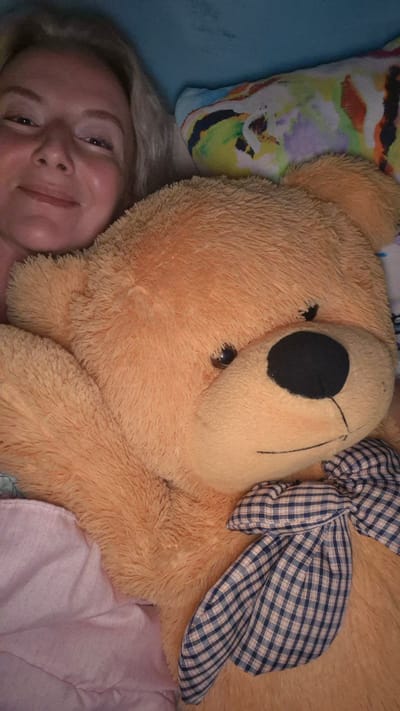 The image shows a close-up of a woman lying in bed with a large teddy bear. The woman is on the left side of the frame, her face is partially visible, and she is smiling. She has blonde hair and appears to be wearing makeup. The teddy bear is large and fluffy, with orange fur, a black nose, and a plaid bow tie. The background is blurred and appears to be a bed with pillows. There is no indication of the location being Mynbaev, Kazakhstan.