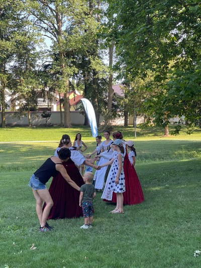 A group of people, including adults and children, are gathered in a grassy park area. Several women are dressed in traditional or festive attire, with long skirts and white blouses. One woman in a black tank top and denim shorts is interacting with a young boy wearing a green t-shirt and patterned shorts. Another woman, wearing a long red skirt, appears to be leading or participating in a dance or activity with others. A tall, thin, blue object, possibly a flag or banner, stands in the background. Trees and buildings are visible behind the people, suggesting an outdoor public space. The overall scene depicts a communal gathering or performance in a park-like setting. This location is the park in the city of Stupava, Slovakia.