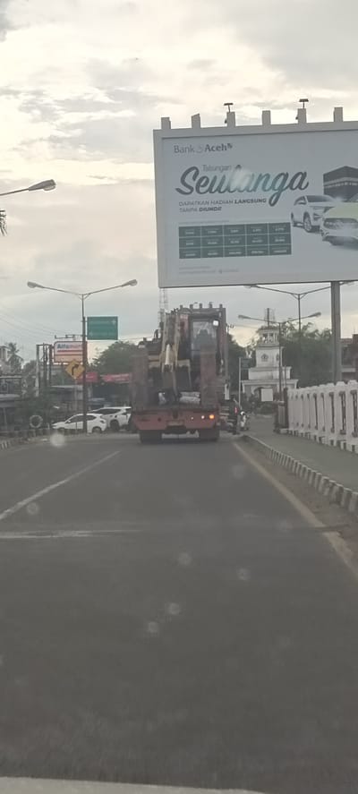 The image captures a road scene from a vehicle, with a large flatbed truck carrying an excavator in the immediate foreground, moving away from the viewer. The road appears damp or reflective, possibly due to rain or light conditions, and there are visible specks on what could be a windshield or camera lens.
Above the road, under an overcast sky, a large billboard prominently displays an advertisement for "Bank Aceh," featuring "Tabungan Seulanga" (Seulanga Savings). The billboard promises "DAPATKAN HADIAH LANGSUNG TANPA DIUNDI!!" (GET DIRECT PRIZES WITHOUT A DRAW!!) and includes a table with various monetary values and images of cars, likely as prizes. The bank's website, "www.bankaceh.co.id," is also visible.
To the left, streetlights illuminate the area, and a green road sign with text such as "WEATANG RAYA" provides directional information. Below it, a yellow road sign indicates a curve. An "Alfamart" convenience store sign can be seen further down the street. Several other vehicles, including white cars, are visible in traffic. On the right side of the road, a white decorative barrier lines the street, and a distinctive white architectural structure resembling a mosque with a clock tower is visible in the distance. The scene suggests a cloudy daytime setting in an urban or semi-urban area, consistent with Dayahlhoksukon Barat, Indonesia.