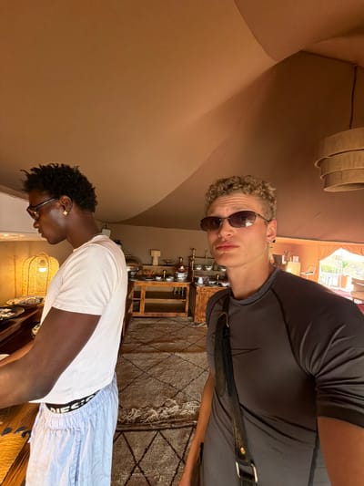 Two young men are photographed in a large, tent-like structure with a brown canvas ceiling in Agafay, Morocco. The man on the left, wearing a white t-shirt and light blue pajama pants with the waistband "DIESEL" visible, has dark skin, short curly dark hair, and is wearing dark sunglasses and a gold earring. He is looking to his left. The man on the right, with lighter skin and short, curly blonde hair, is wearing a dark gray t-shirt and sunglasses. He is looking directly at the camera and has a black strap with gold hardware slung across his chest. In the background, there is a wooden buffet table laden with various dishes, bowls, and serving utensils. A woven basket lamp is visible to the left of the buffet. The floor is covered with a patterned rug. The overall impression is that of an indoor dining area within a desert camp or resort.