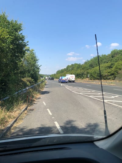 A sunny day with a clear blue sky over a highway in Enborne Row, United Kingdom. The road is multi-lane and appears to be heading into a slight bend. On the right side of the road, dense green trees line the embankment. In the foreground, a car's dashboard and windshield are visible, with an antenna casting a shadow. Further down the road, there are several vehicles, including vans and cars, with some appearing to be moving in a queue. One van prominently displays the logo "PIMM'S". The left side of the road features a metal guardrail and overgrown vegetation.